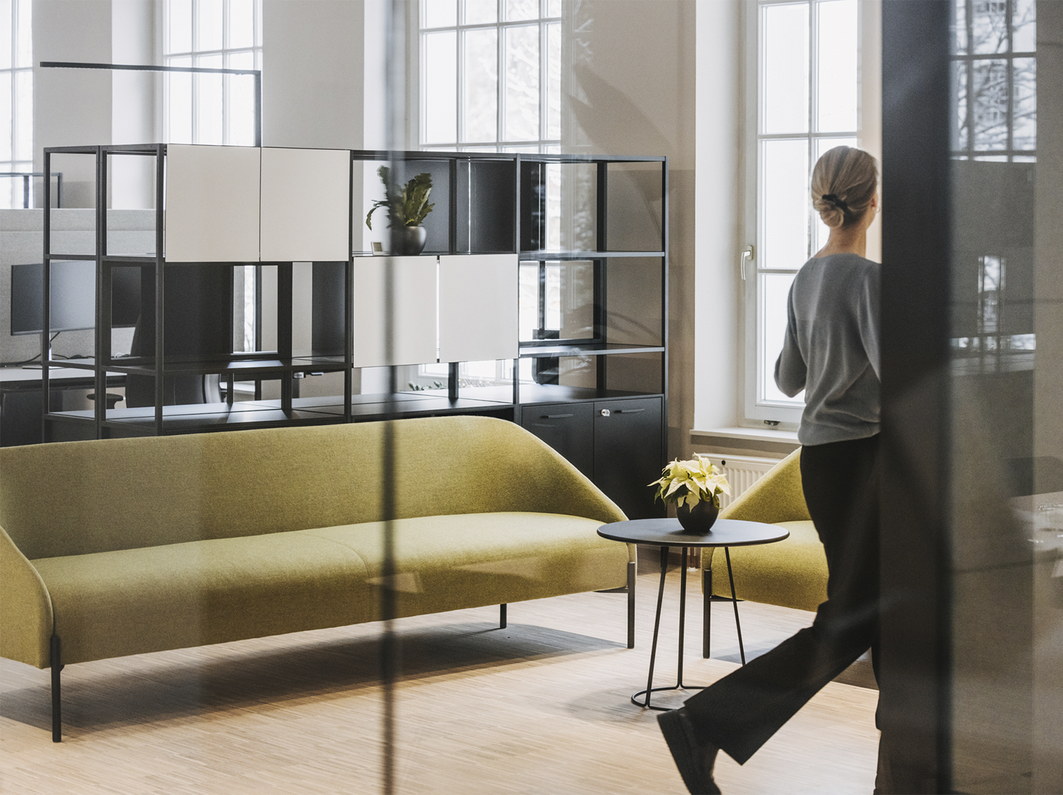 Modern office lounge with a green sofa, black table with a plant, and a woman walking past large windows.